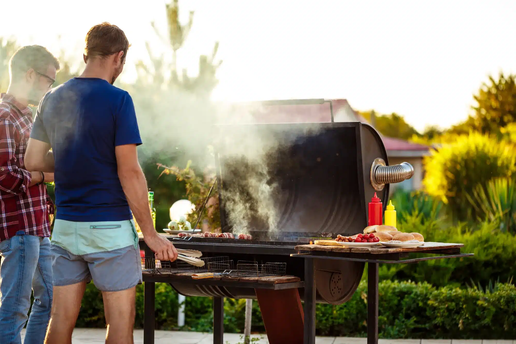 Backyard barbecue with a smoker in action, two men grilling food and preparing flavorful smoker recipes for a summer feast.