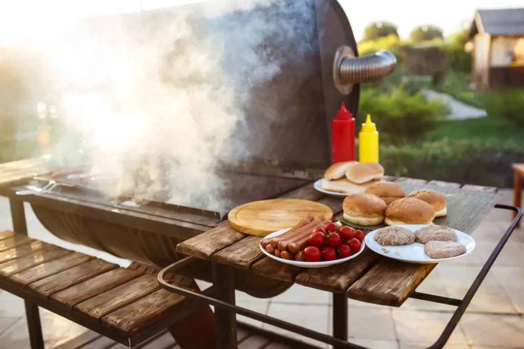 Smoker in a backyard with smoke rising and a wooden table set with burger buns, sausages, fresh tomatoes, and condiments.