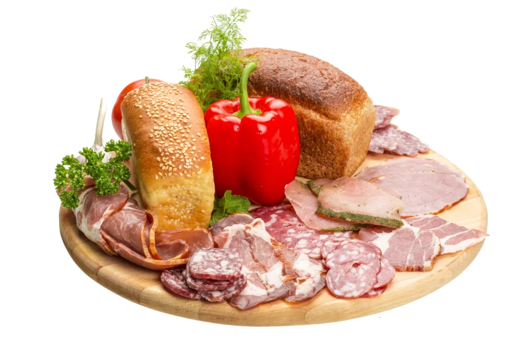 Assorted lunch meats with artisan bread and fresh vegetables on a wooden board.