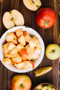 Chopped apples in a white bowl surrounded by whole and halved apples on a wooden surface.