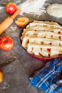 nbaked apple tart in a red dish with lattice pastry, surrounded by fresh apples, rolling pin, and flour on a rustic kitchen countertop