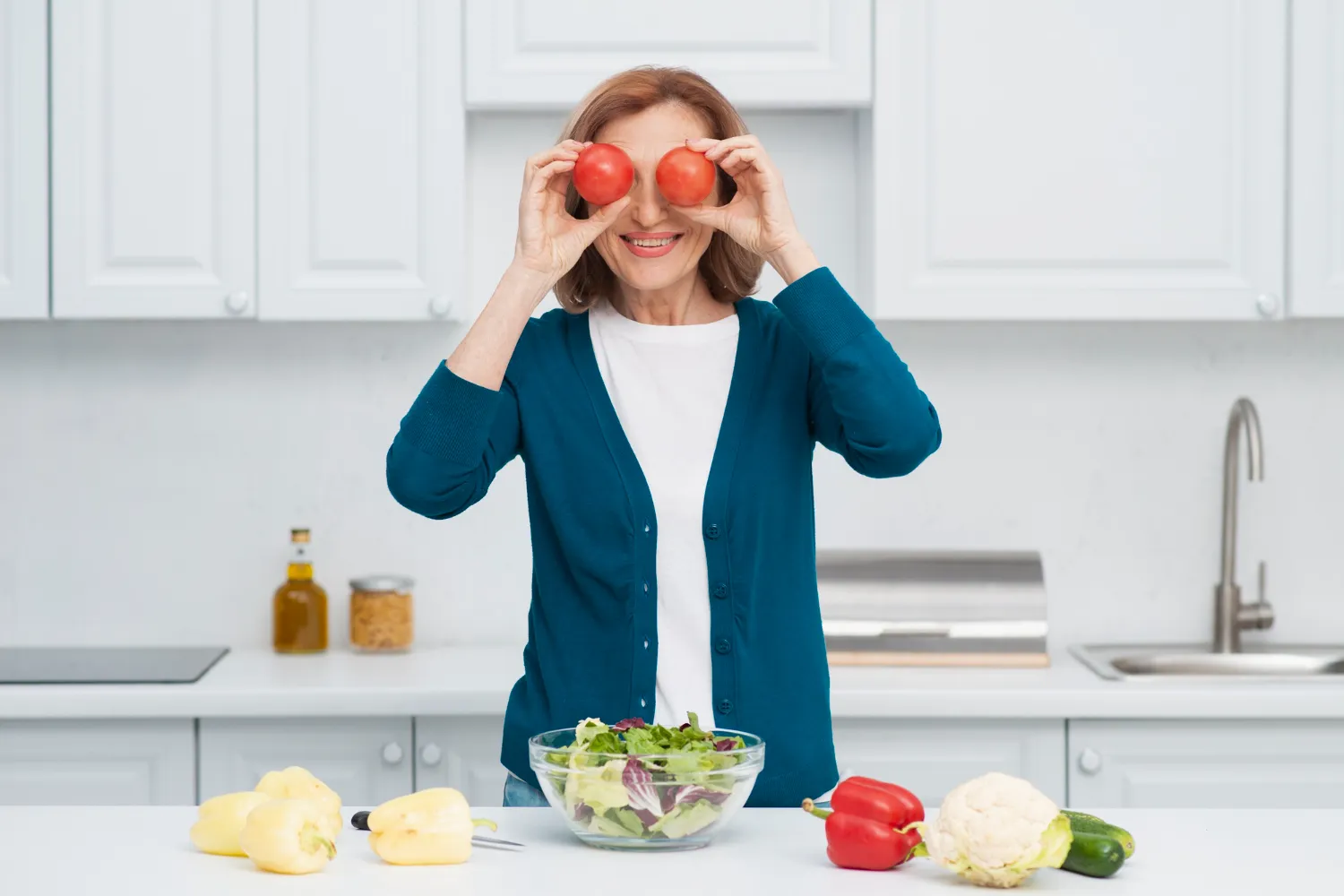 A cheerful woman holding two tomatoes over her eyes, surrounded by fresh vegetables and a salad bowl, representing the menopause diet 5-day plan.