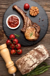 Grilled tomahawk steak with a side of tomato sauce, fresh cherry tomatoes, a loaf of artisan bread, and roasted garlic on a rustic wooden table.