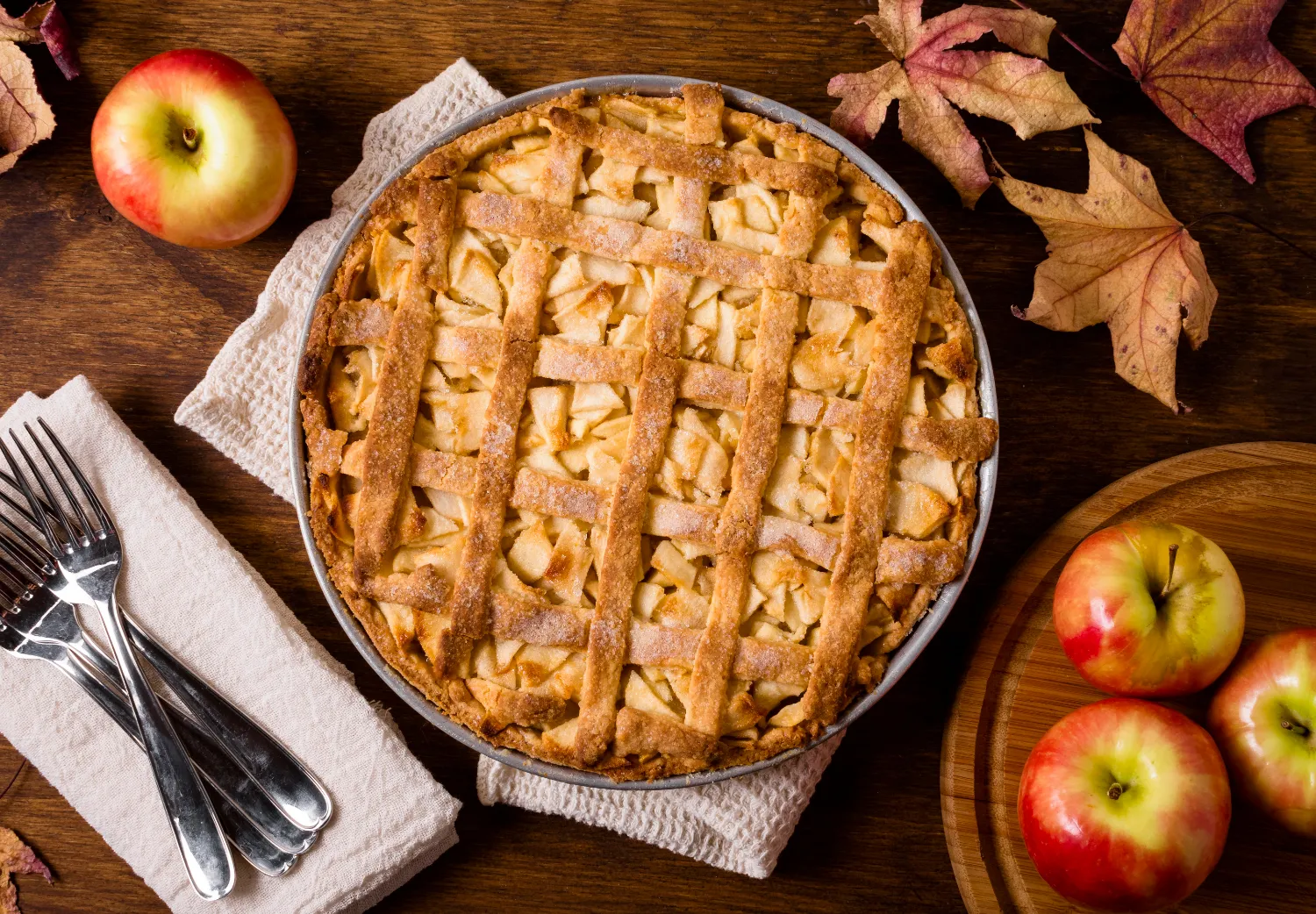 Freshly baked apple and pecan tart with a golden lattice crust, surrounded by apples and fall leaves on a rustic wooden table.