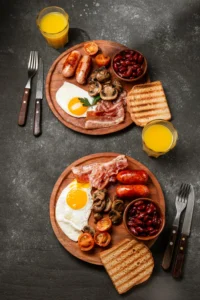 Full English breakfast with eggs, bacon, sausages, mushrooms, beans, toast, and orange juice on a dark table.