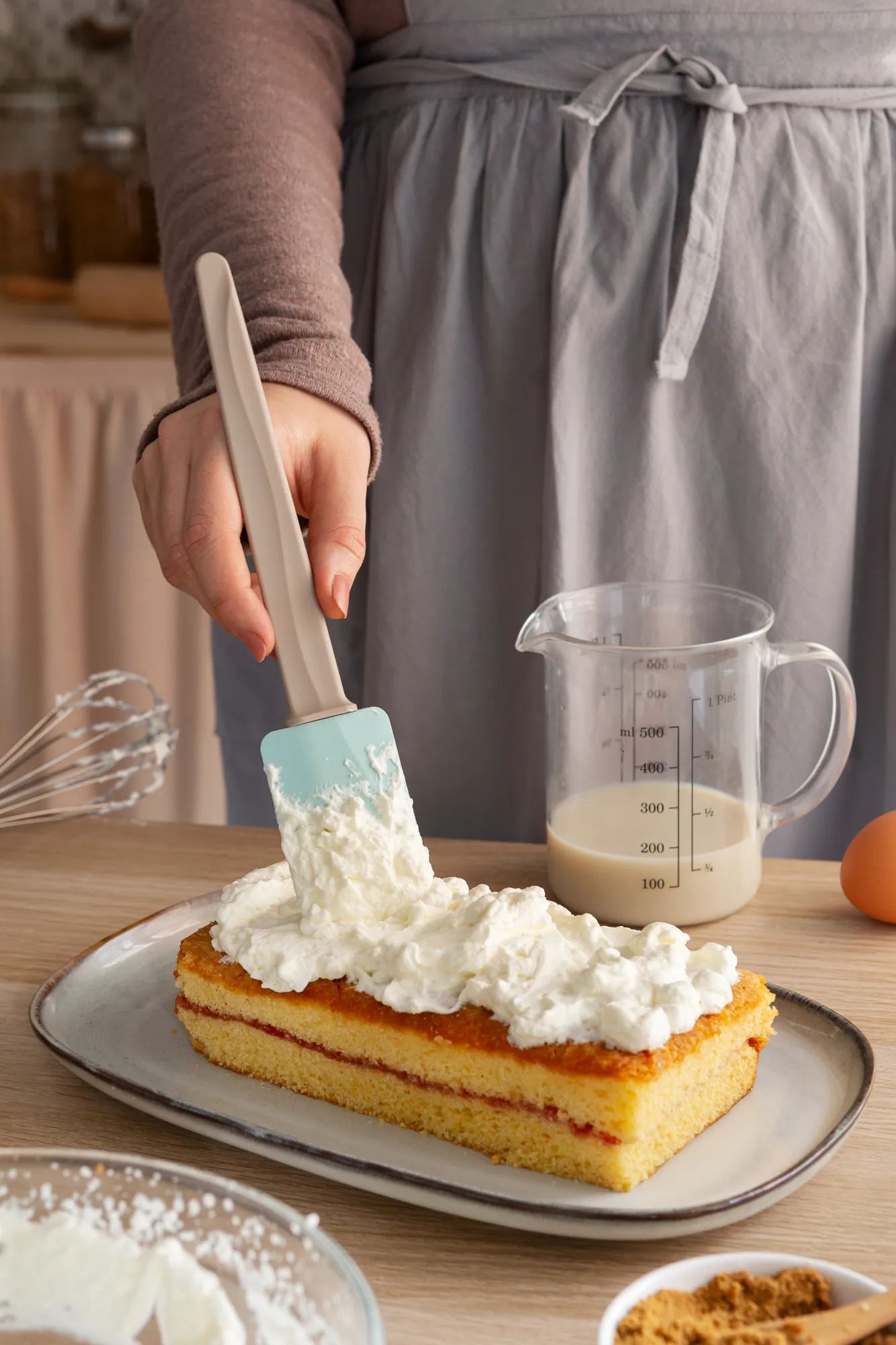 Applying whipped cream with a spatula on a layered kefir sheet cake with jam filling, placed on a serving platter.