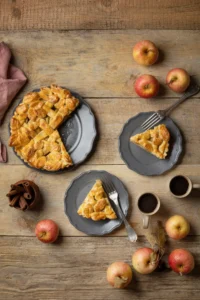 Slices of apple tart on gray plates with fresh apples and coffee cups on a rustic wooden table.