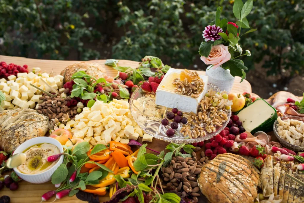 An assortment of sorghum festival food, featuring bread, cheeses, fruits, nuts, and a decorative floral arrangement on a rustic wooden table.