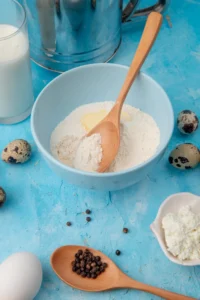 Ingredients for baking kefir sheet cake, including flour, milk, eggs, and butter in a blue mixing bowl with a wooden spoon on a vibrant blue background.