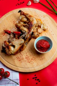 Crispy rotisserie chicken served with roasted potatoes, garlic, chili peppers, and a bowl of tomato sauce on a wooden platter.