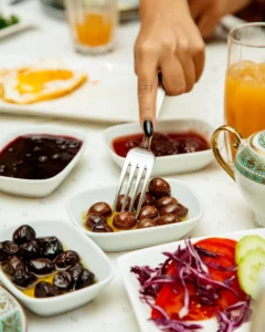 A hand with a fork picking olives from a small white dish, surrounded by various colorful side dishes, including fresh vegetables, jams, and beverages, on a breakfast table.