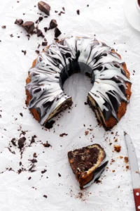 Chocolate cream cheese pound cake with a marble glaze, partially sliced, surrounded by chocolate chunks on a white textured surface