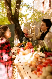 A vendor and a customer interacting at a farmers' market, with fresh produce like apples, radishes, and squash displayed on a wooden stand.