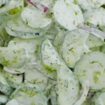 A close-up of creamy cucumber salad with thinly sliced cucumbers, red onions, fresh dill, and a creamy dressing topped with black pepper.