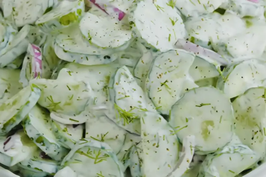 A close-up of creamy cucumber salad with thinly sliced cucumbers, red onions, fresh dill, and a creamy dressing topped with black pepper.