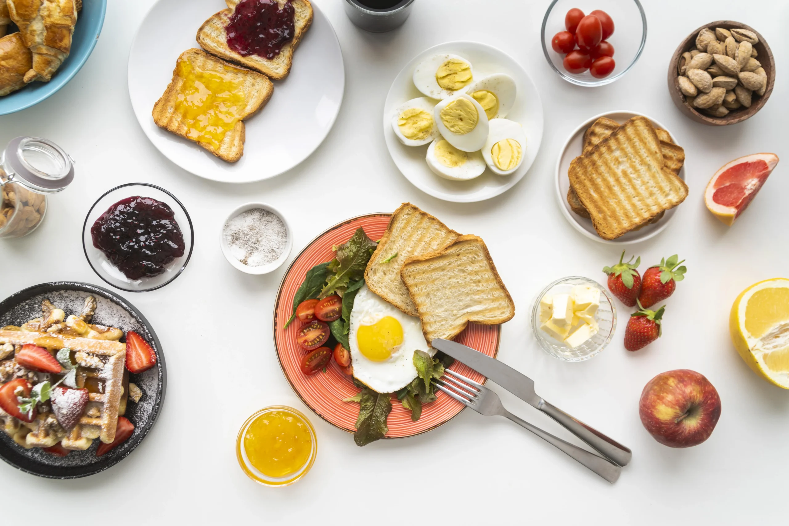 A colorful breakfast table with toast, eggs, fruit, jam, waffles, and fresh vegetables, offering a variety of healthy breakfast sides.