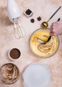 Hand mixing cake batter with a spatula in a glass bowl, surrounded by cocoa powder, a stand mixer, and spices on a beige countertop.