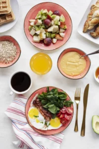 Breakfast sides featuring eggs, bacon, greens, fresh fruit salad, oatmeal, croissants, orange juice, and coffee for a healthy morning meal.