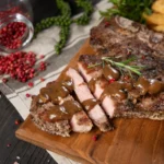 Grilled beef brisket with peppercorn sauce served on a wooden board with crinkle-cut fries and fresh herbs.