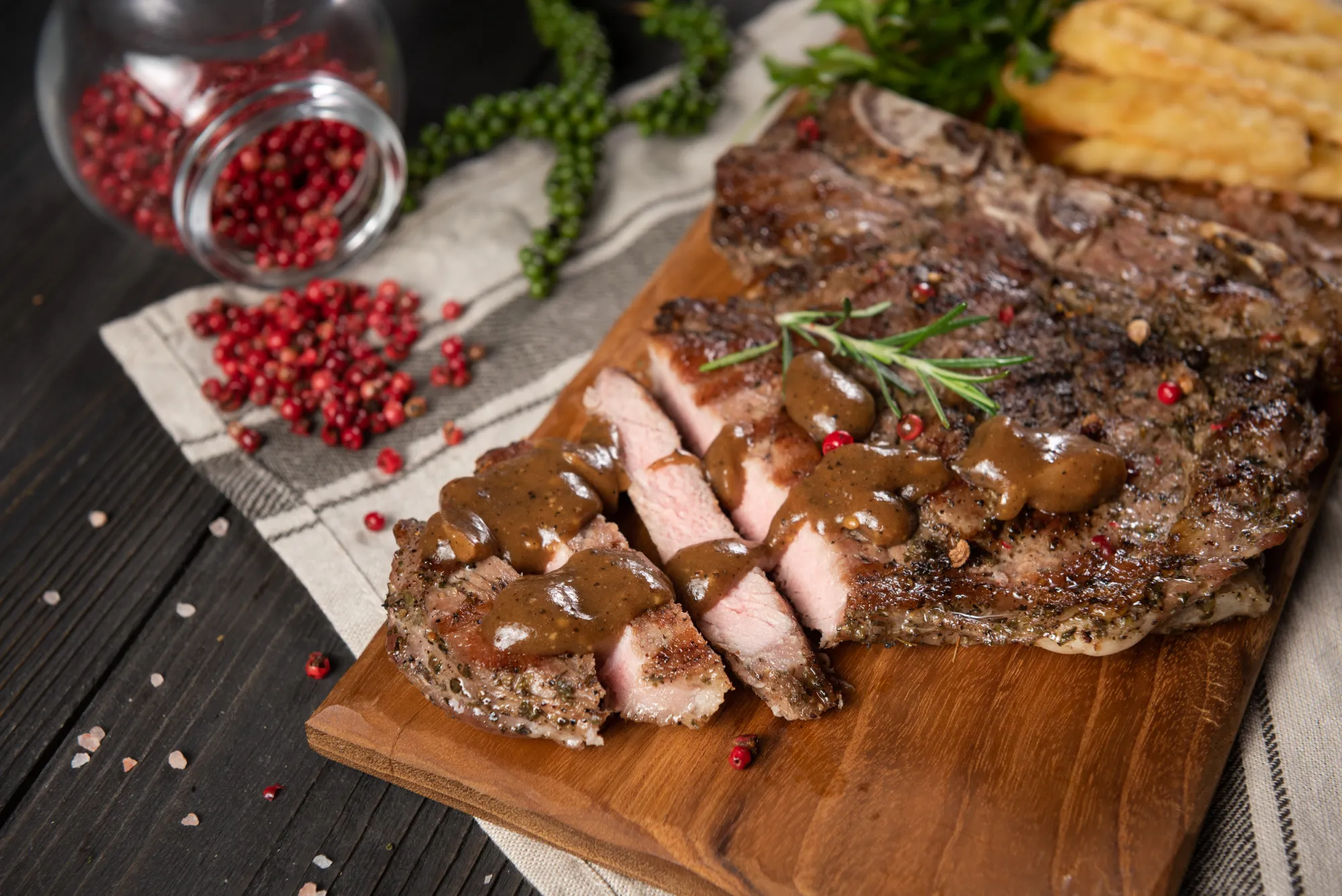 Grilled beef brisket with peppercorn sauce served on a wooden board with crinkle-cut fries and fresh herbs.