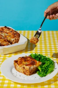 A slice of hotdish resembling the Rick Nolan Hotdish Recipe, served on a white plate with fresh green lettuce on the side, placed on a yellow gingham tablecloth. A hand holds a fork with a piece of the dish against a bright blue background