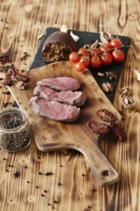 Slices of fresh, tender beef brisket on a wooden cutting board with cherry tomatoes, bread, black pepper, dried tomatoes, and walnuts.