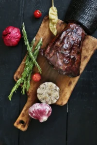 Texas-style smoked beef brisket in slices on a wood chopping board with garlic, rosemary sprigs, cherry tomatoes and roasted peppers, ideal for a texas brisket bacon recipe