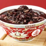 Purple black bean soup served in a decorative red bowl on a bamboo tray with a red background