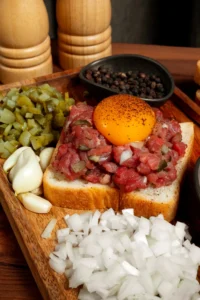 A fresh breakfast featuring corned beef tartare on toast, topped with a raw egg yolk, surrounded by chopped onions, pickles, garlic, and peppercorns.