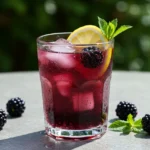 A refreshing glass of blackberry lemonade summer drink with lemon slice, blackberry, and mint garnish on a sunny outdoor table.
