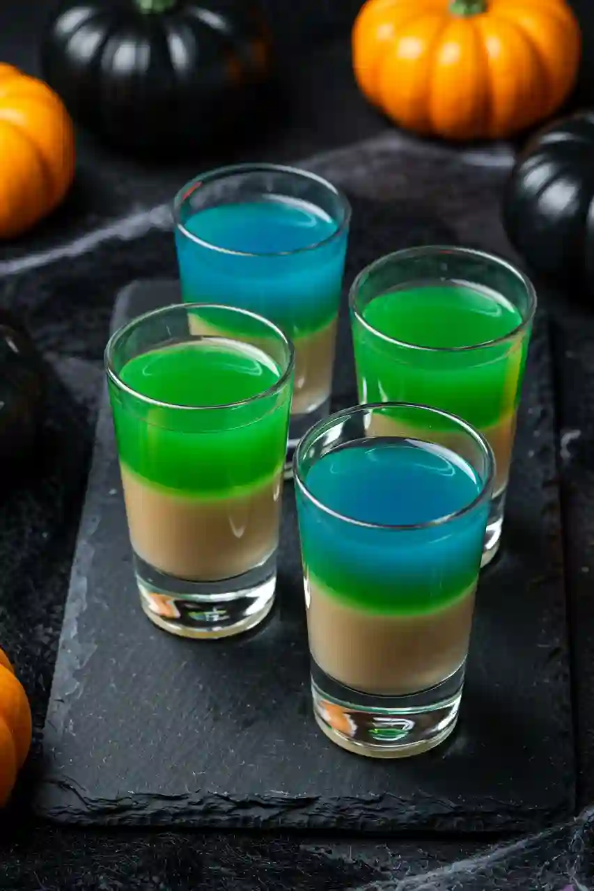 Boogie Man Halloween Shot Four Boogie Man layered shots with beige, green, and blue colors in clear shot glasses, styled with pumpkins and cobwebs on a black slate.