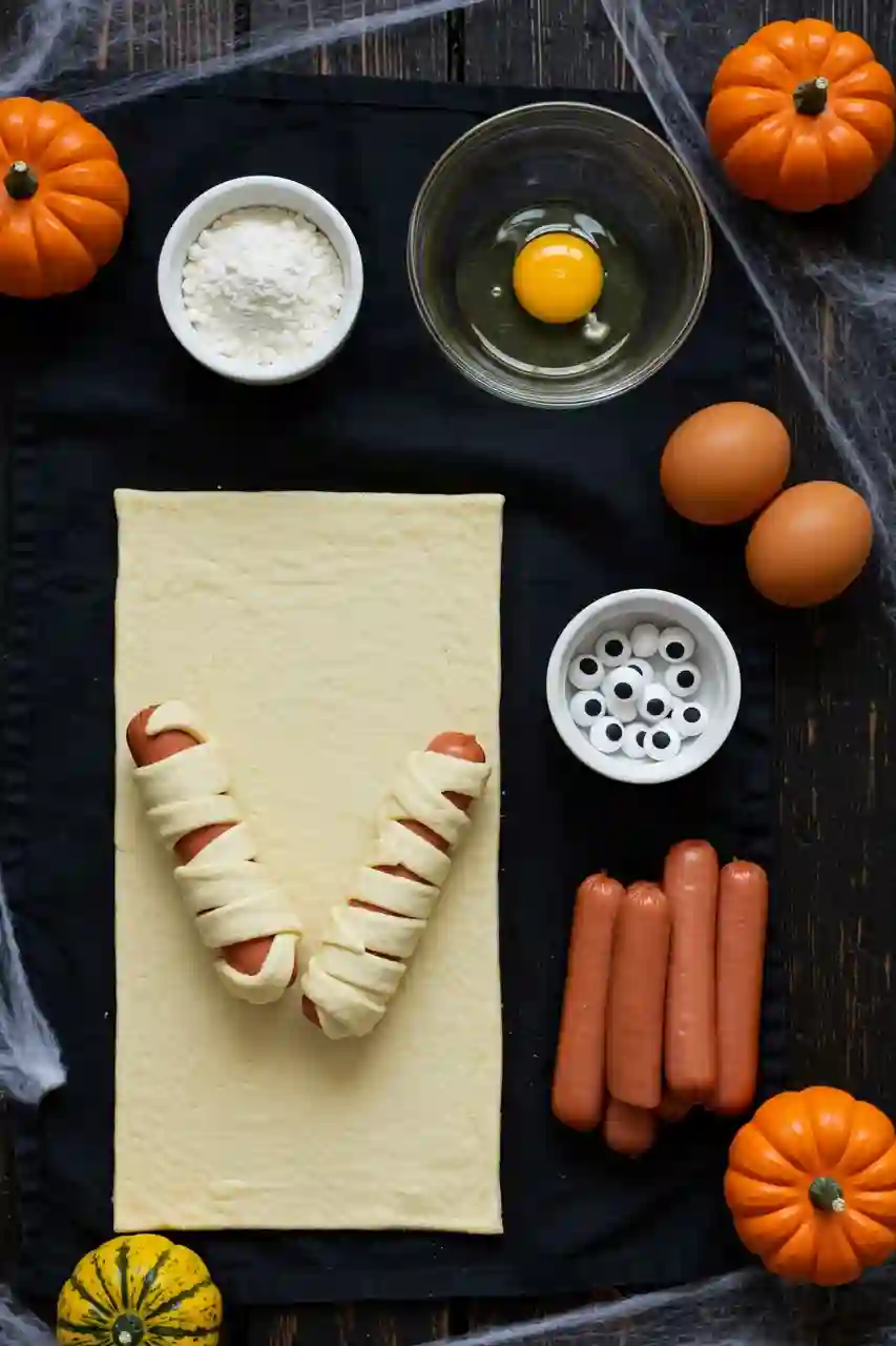 halloween-mummy-hot-dogs-ingredients Ingredients for Halloween Mummy Hot Dogs including puff pastry, hot dogs, mustard, egg, and candy eyes on a wooden table.